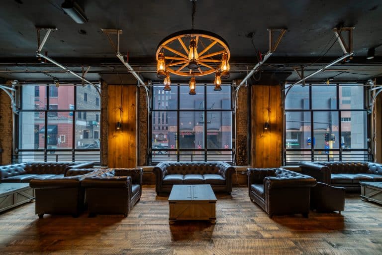 A stylish lounge with leather sofas, barnwood flooring, large windows, and a wagon wheel chandelier overlooking city buildings. The space boasts an industrial and rustic design.