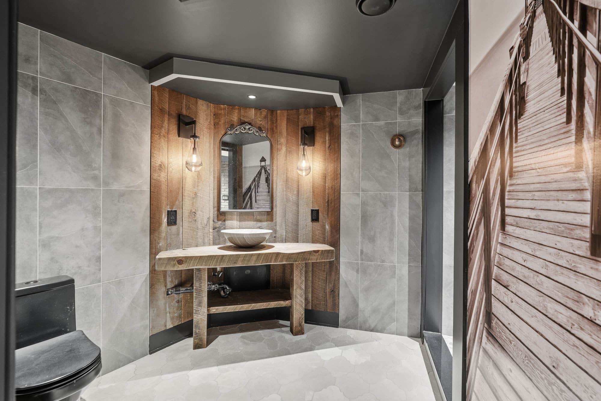 A modern bathroom with gray tiled walls, a rustic wood vanity, a round vessel sink, a decorative mirror, and two wall-mounted lights. A black toilet and a large photo of a wooden boardwalk are also visible.